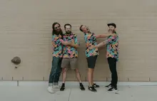 The four members of Atomicos are hugging while standing in front of a beige brick wall. The bass player does not appear to be enjoying himself.  